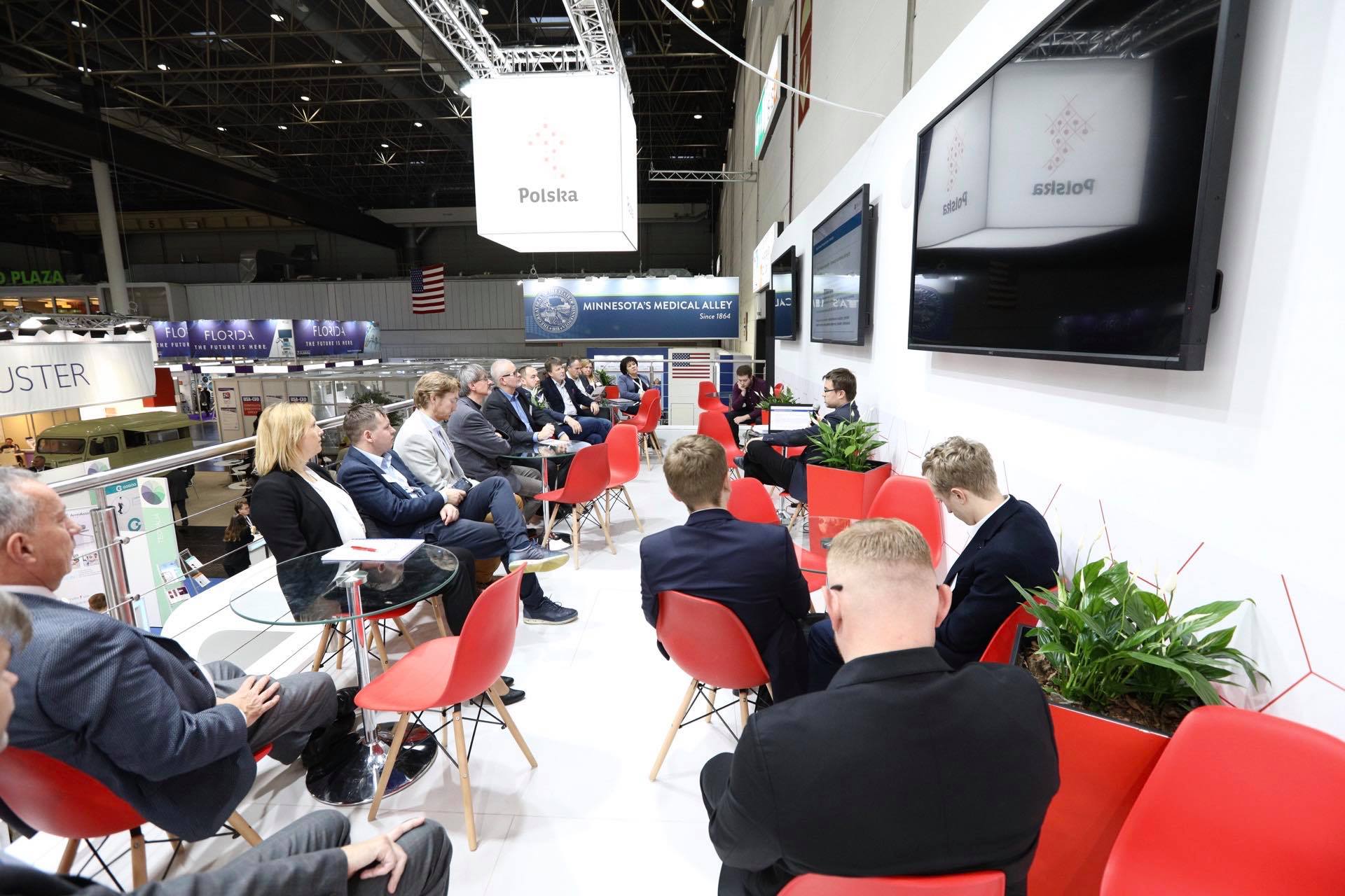 grupa ludzi w różnym wieku siedzi na czerwonych krzesełkach na antresoli. W tle widać trzy ekrany wiszące na ścianie. Z sufitu zaś zwisa sześcian z napisem Polska