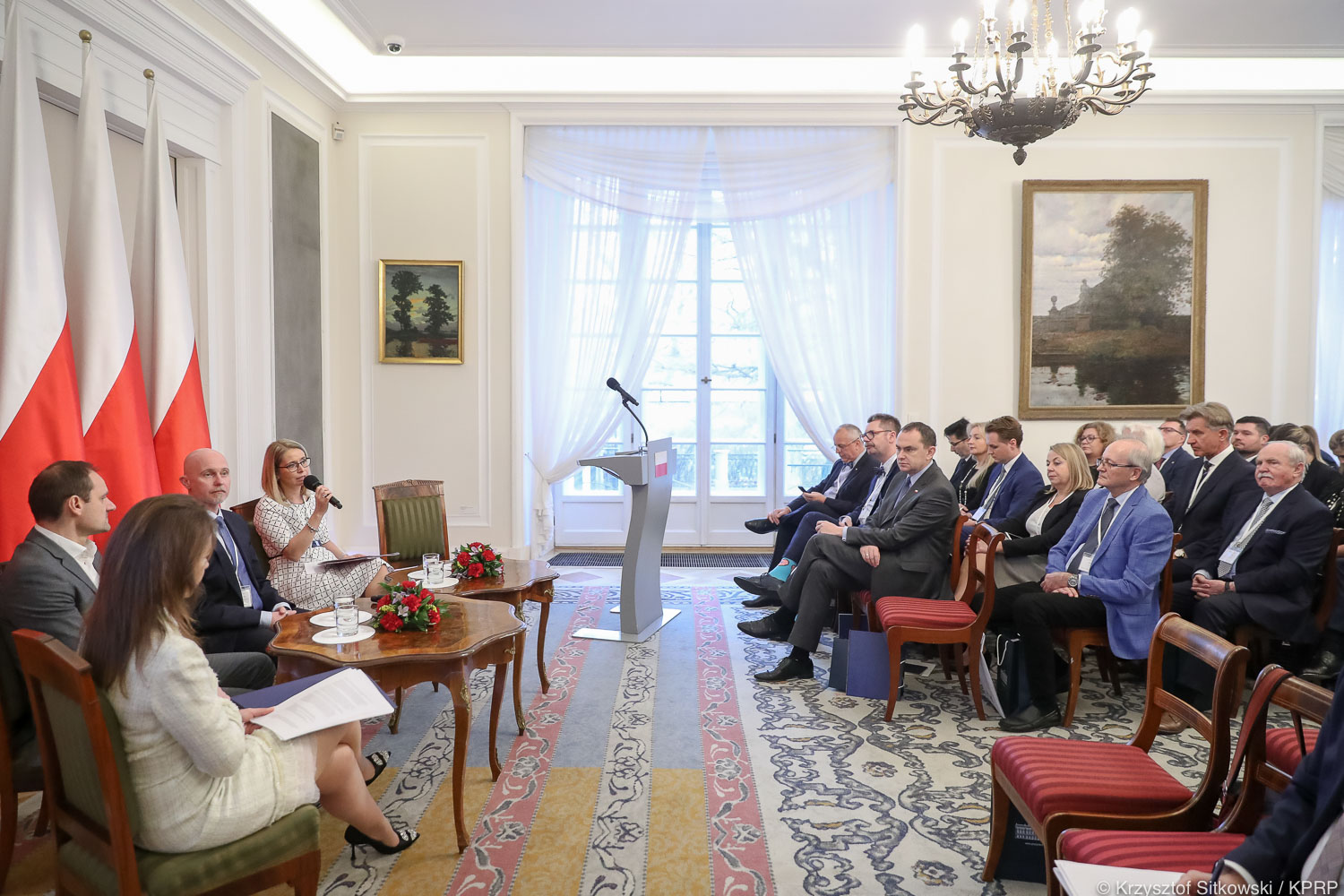 Na zdjęciu widać uczestników konferencji - panelistów siedzących na krzesłach po lewej stronie i uczestników siedzących na krzesłach po prawej stronie