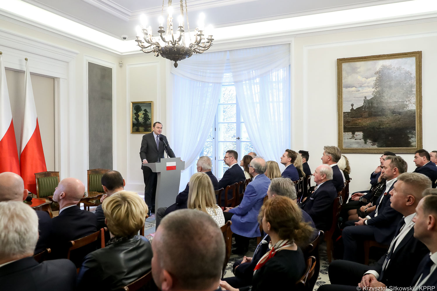 Na zdjęciu widać flagę polski, a przy niej mównicę, przy której stoi jeden z panelistów konferencji, na pierwszym planie widać uczestników konferencji siedzących na krzesłach.
