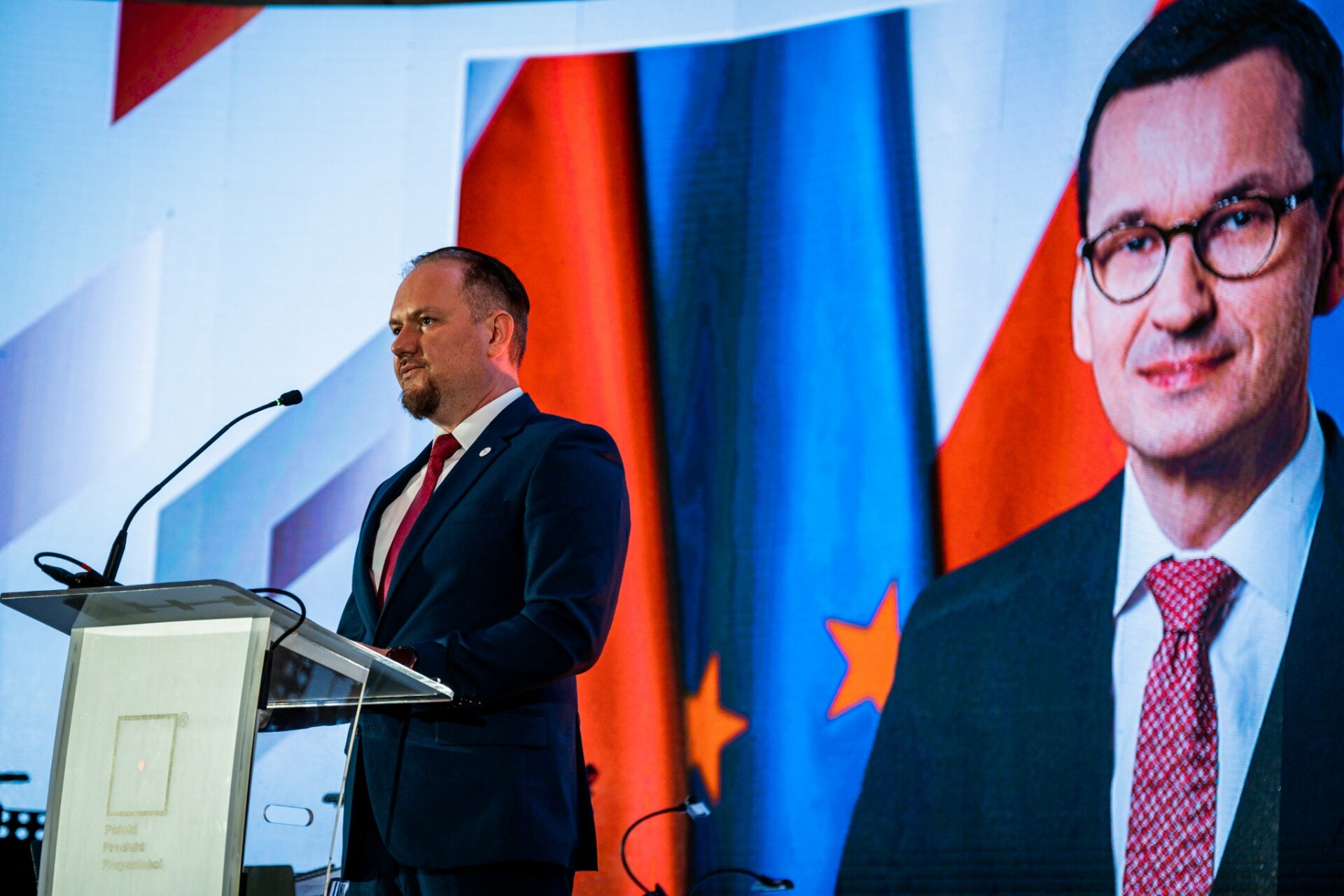 Dariusz Budrowski, prezes PARP przemawia na tle zdjęcia Mateusza Morawieckiego