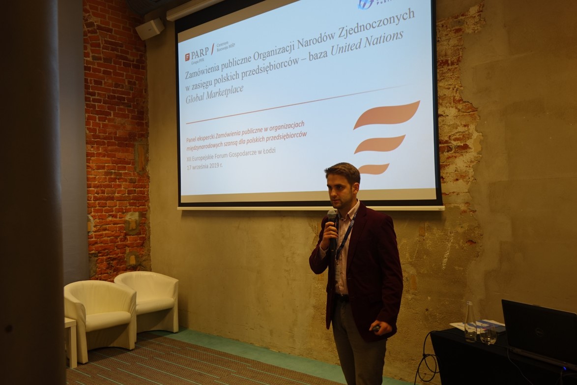 Młody mężczyzna stoi na sali z mikrofonem w ręku. Za nim na ścianie jest umieszczony ekran. Jest na nim widoczny napis „Zamówienia publiczne Organizacji Narodów Zjednoczonych w zasięgu polskich przedsiębiorców – baza Unites Nations Global Market”.