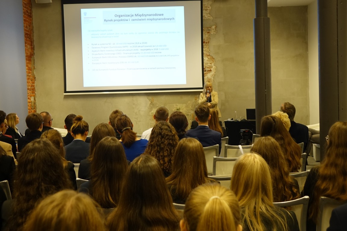 ludzie siedzą na sali. Na jej końcu widać podium, na którym stoi kobieta. Za nią widać ekran