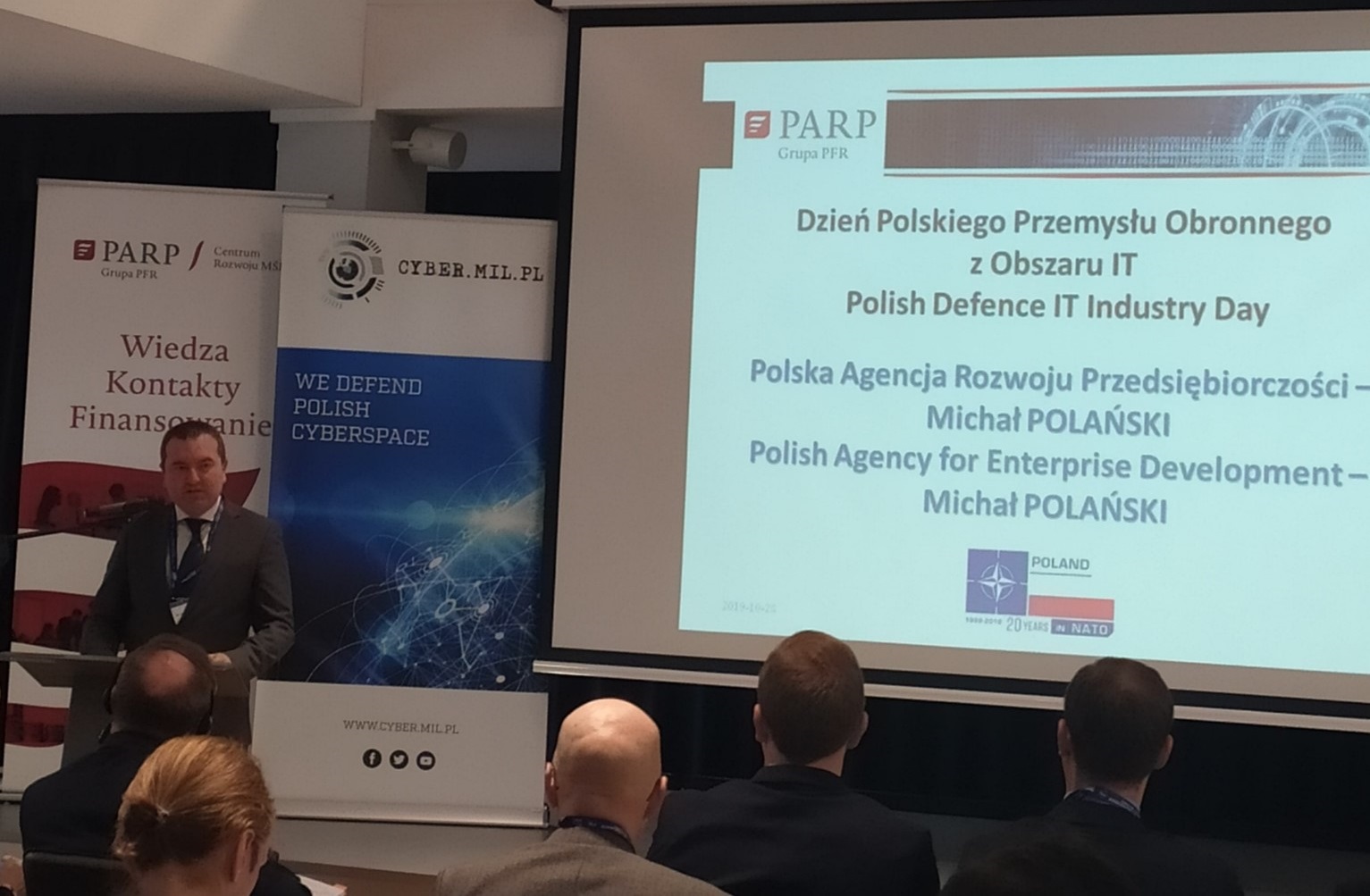 Na pierwszym planie po prawej stronie widać mężczyznę w średnim wieku, który stoi przy mównicy. Na lewo od niego znajduje się ekran z wyświetlanymi slajdami prezentacji.