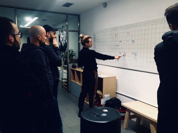 młoda kobieta ubrana w dres stoi przy białej tablicy z tabelkami i wskazuje na jedną z nich trzymanym w ręku flamastrem. Za kobietą stoi trzech mężczyzn w młodym i średnim wieku. Pierwszy z nich ma brodę i czapkę z daszkiem, drugi jest łysy i ma brodę, a trzeci ma krótkie czarne włosy i nosi okulary