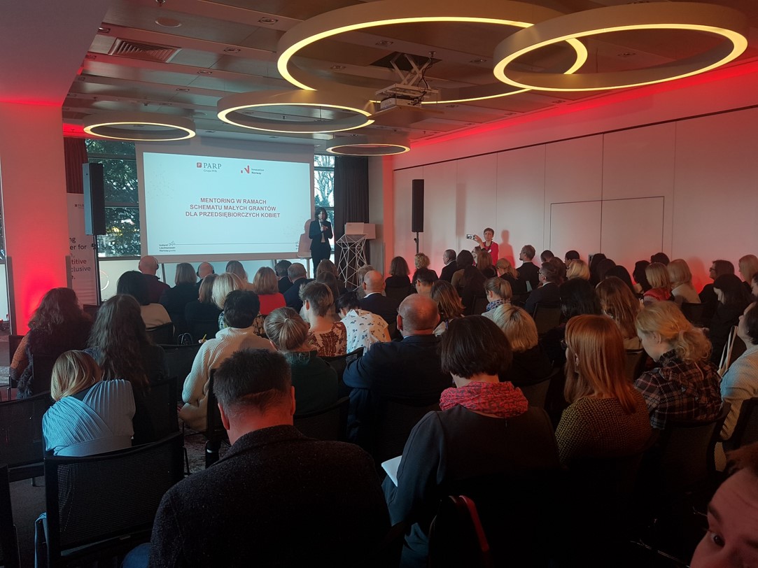 Osoby w różnym wieku, głównie kobiety, siedzą na sali. W głębi widać podest, na którym stoi kobieta. Obok niej widać duży ekran.