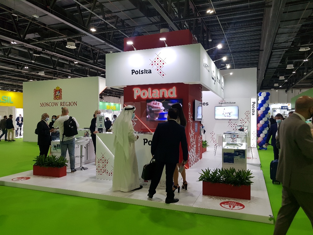 Mężczyzna stoi przez ladą stoiska w przestrzeni wystawienniczej zlokalizowanej w dużej sali. Na...