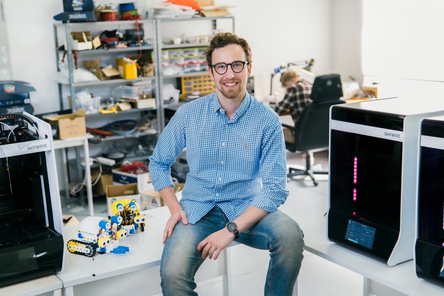 Młody mężczyzna w okularach siedzi w pomieszczeniu, w którym widać drukarki 3D oraz inne przedmioty