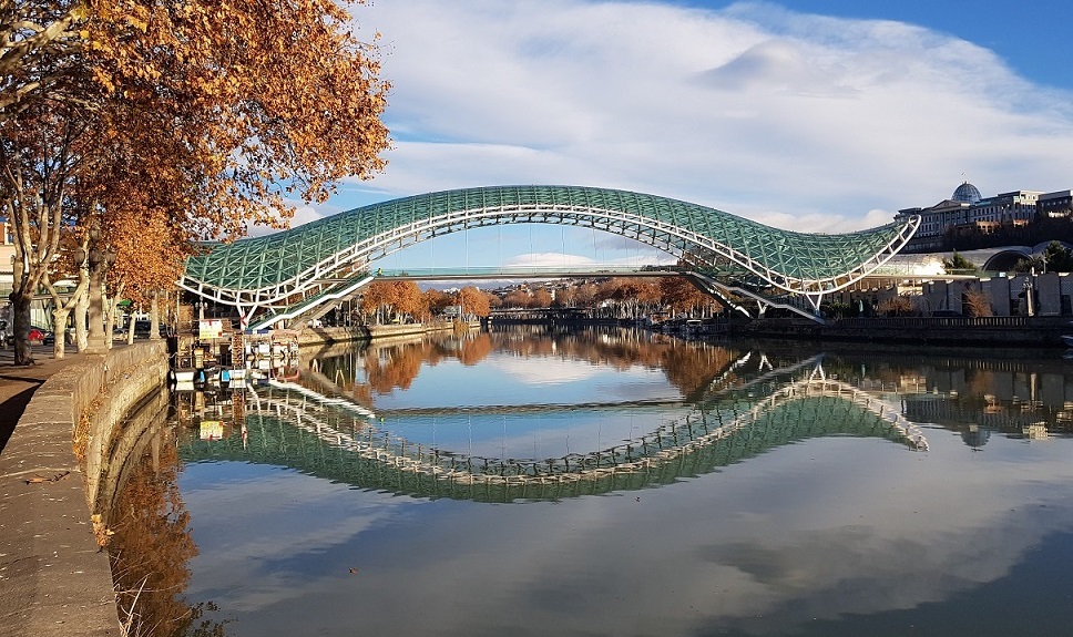 Widok na bulwary miejskie w Tbilisi, na pierwszym planie rzeka, w tle widać most. Po bokach...