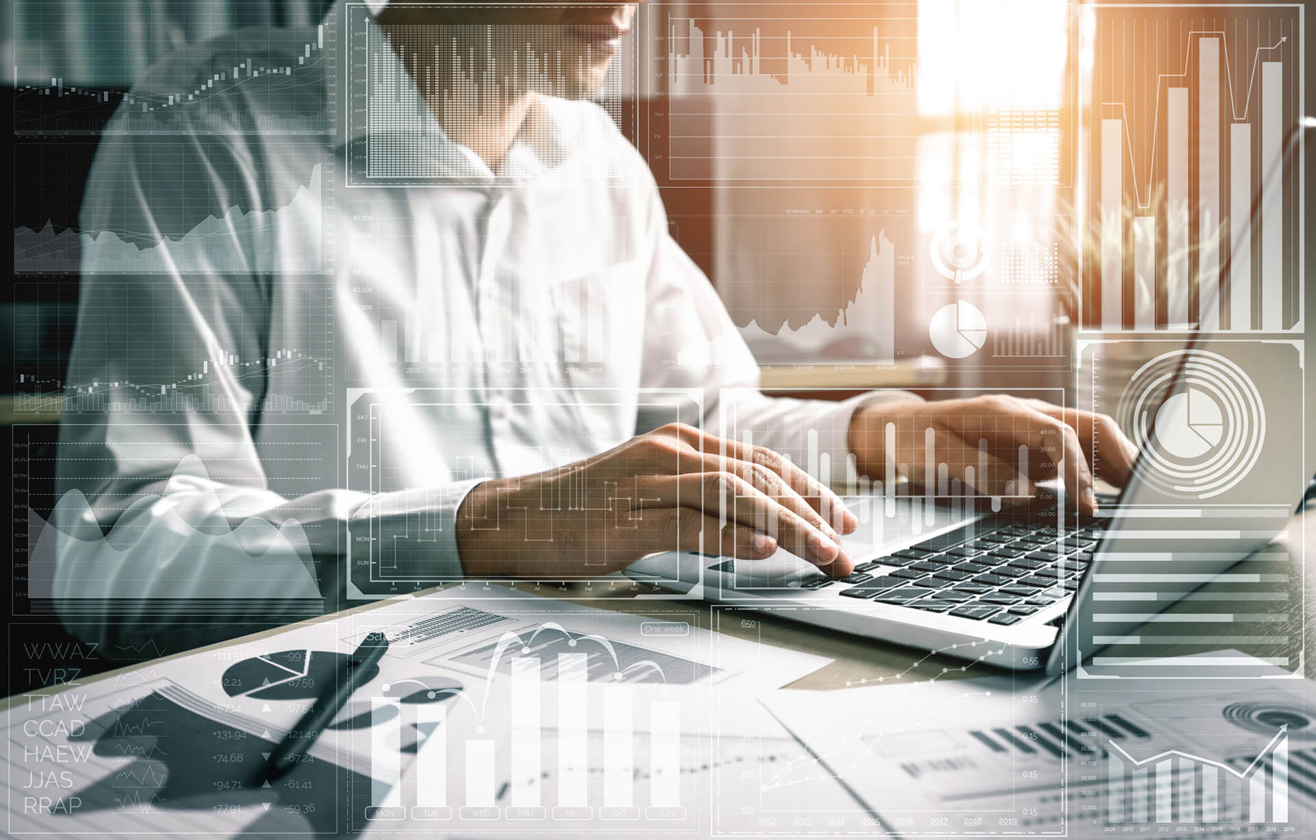 Na zdjęciu widoczny jest mężczyzna w białej koszuli, który pracuje przy komputerze. Na zdjęcie naniesione są grafiki przedstawiające wykresy. 