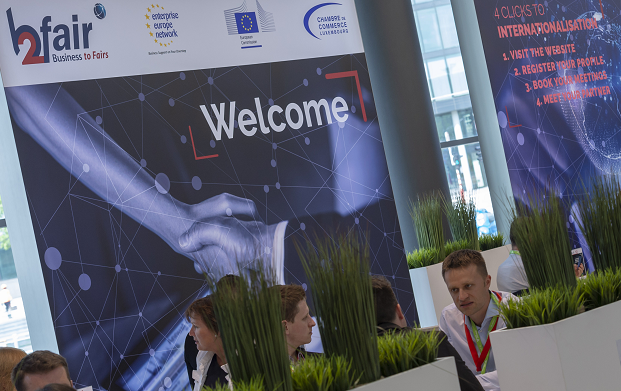 Ludzie siedzą przy stolikach, które są osłonięte wysokimi donicami z roślinami. Na ścianie w tle...