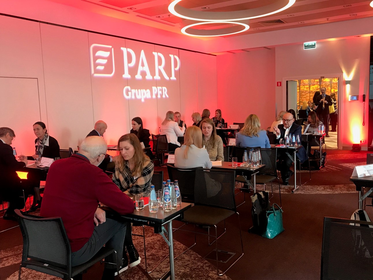 kobiety i mężczyźni w różnym wieku siedzą przy stolikach w sali. Na ścianie widać napis PARP Grupa PFR