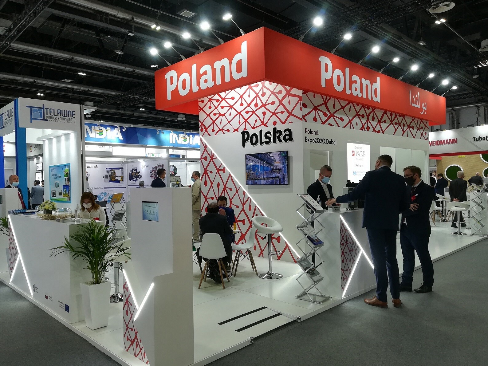 Widok na wnętrze hali wystawowej ze stoiskiem. Na górnej części kiosku widać napis 