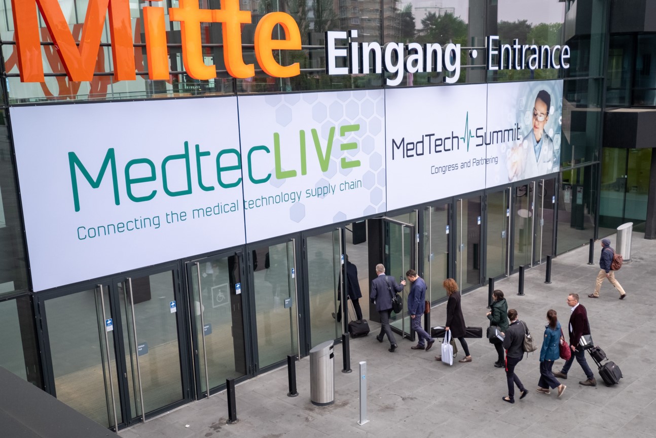 Ludzie wchodzą do nowoczesnego budynku o szklanej elewacji, nad wejściem wisi baner z...