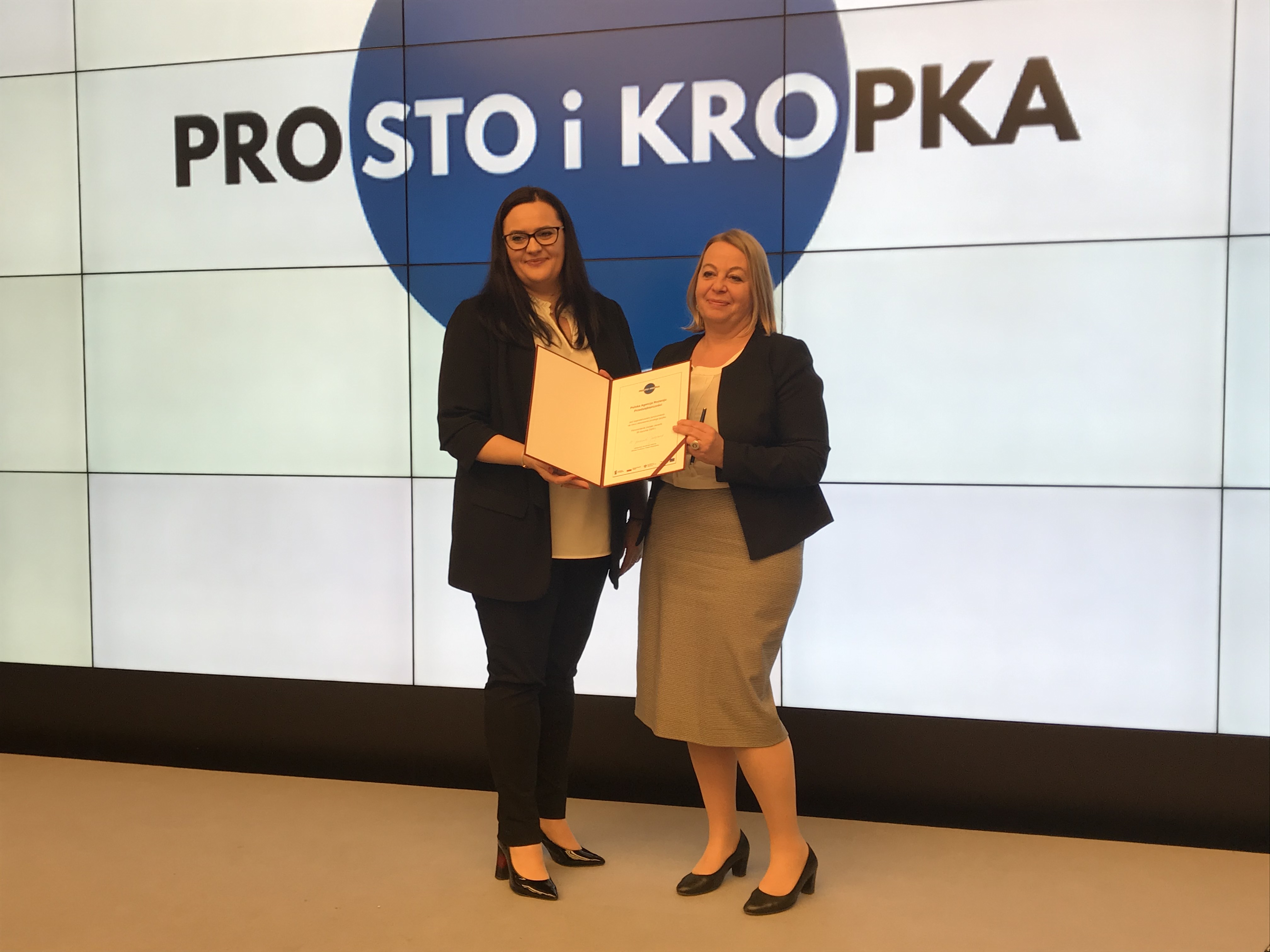 Prezes PARP Małogrzata Oleszczuk stojąca na scenie i wspólnie trzymająca deklarację współpracy na rzecz prostej komunikacji z Minister Funduszy i Polityki Regionalnej Małgorzatą Jarosińską-Jedynak 