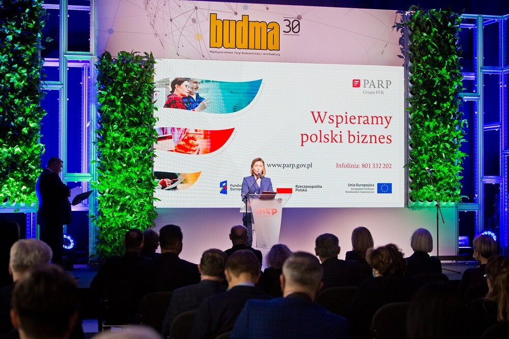 Widzimy uśmiechnięta kobiete w granatowej marynarce, stojącej za białym pulpitem z logo MTP,...