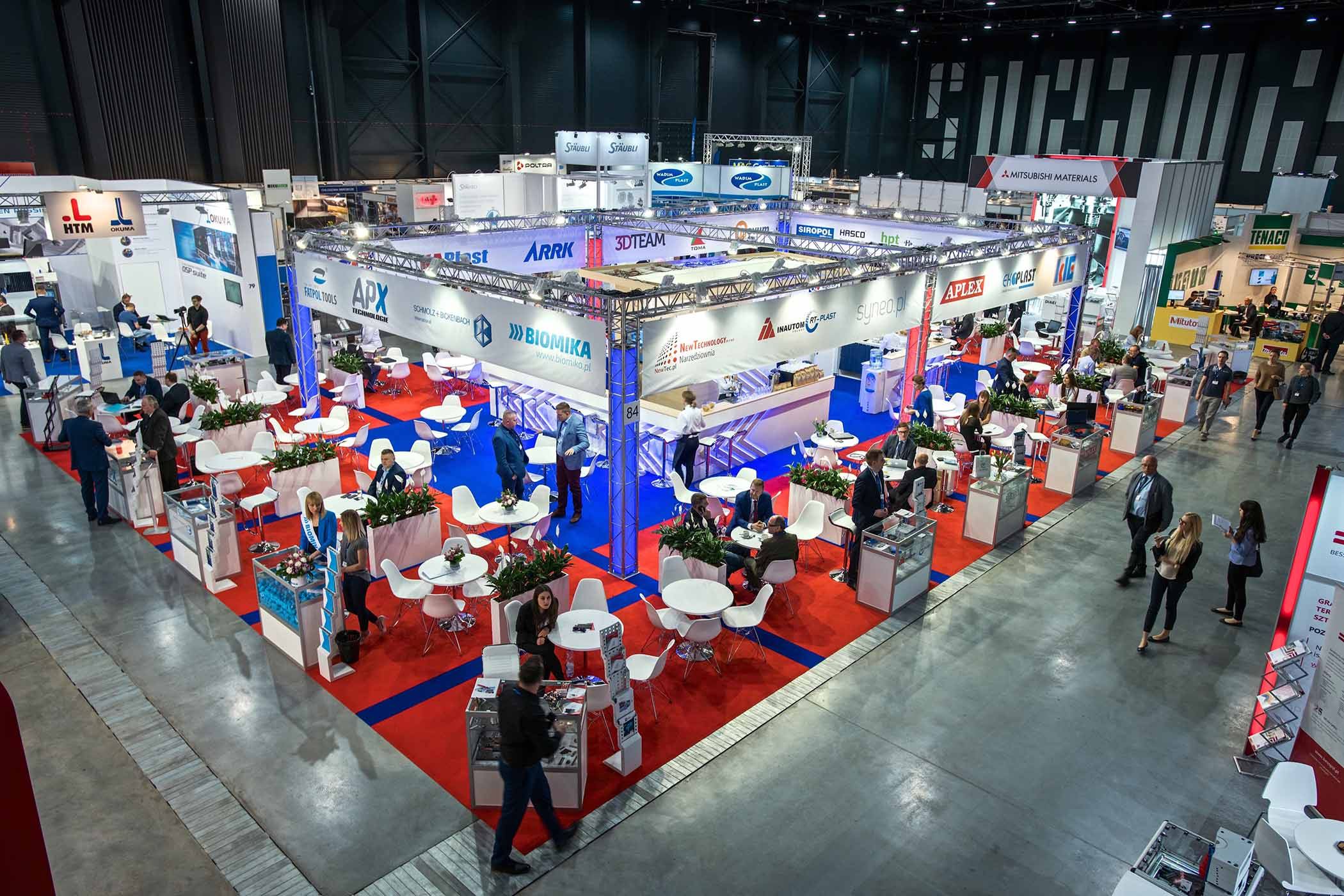 Zdjęcie hali targowej z licznymi stoiskami, mało widoczne w oddali logotypy firm