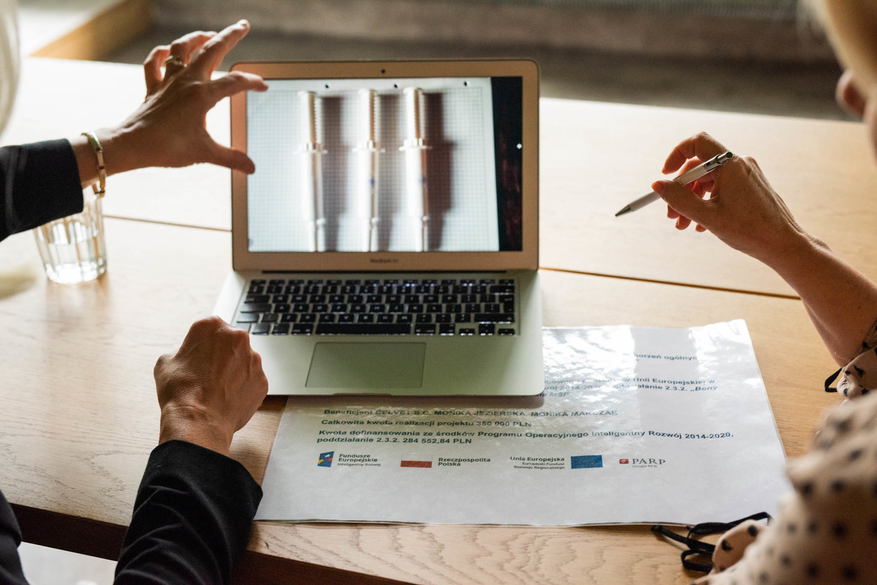 Laptop stoi na biurku, wokół ręce, które wskazują na ekran.